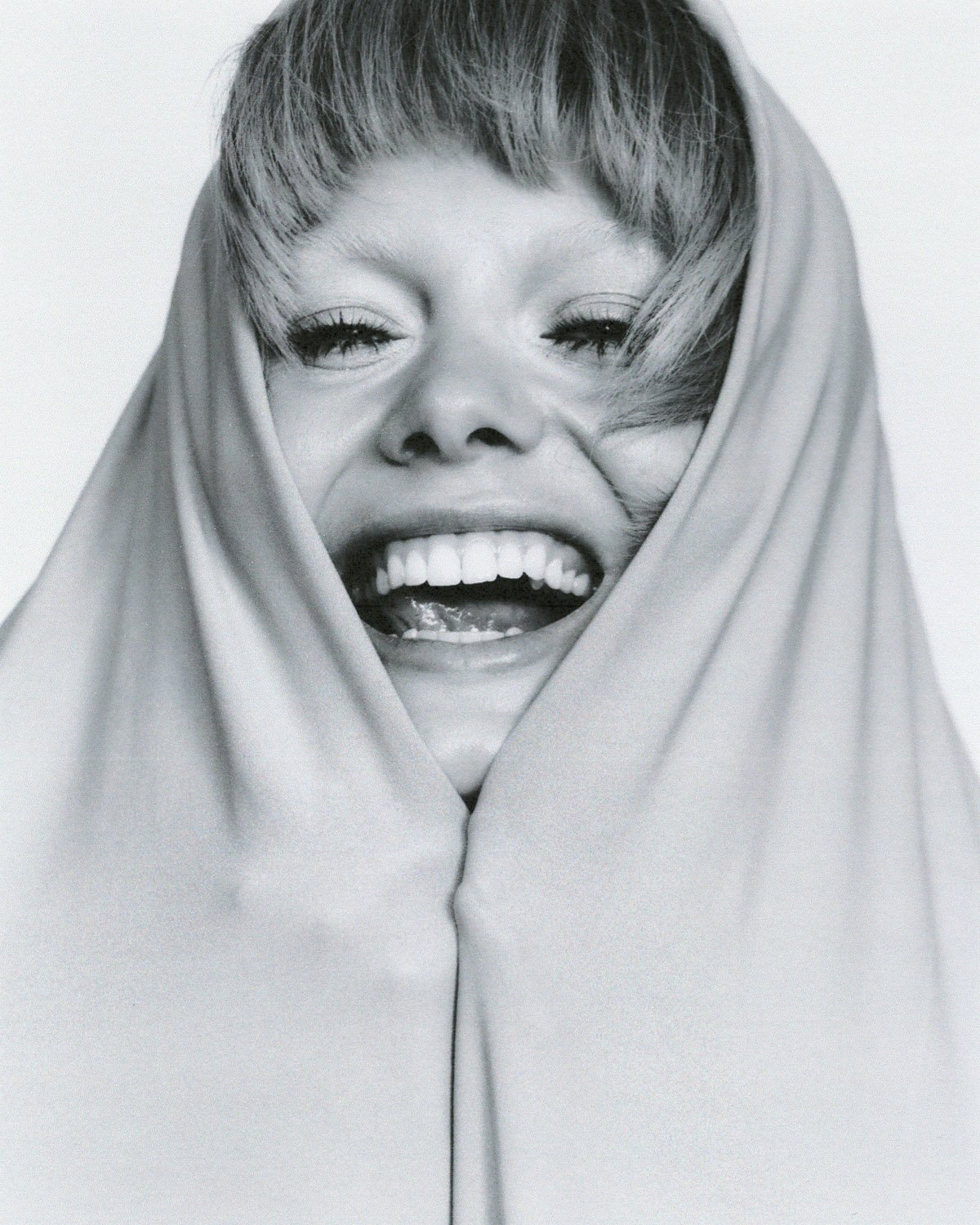 Close-up studio portrait of a smiling woman with fair skin and light blonde hair, draped in a bright white fabric hood.
